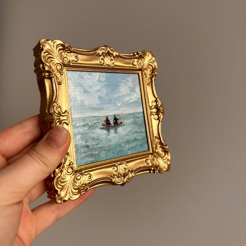 May include: A small, square painting in a gold ornate frame. The artwork depicts two figures in a boat on a turquoise sea under a cloudy blue sky. The frame has a detailed, vintage design.