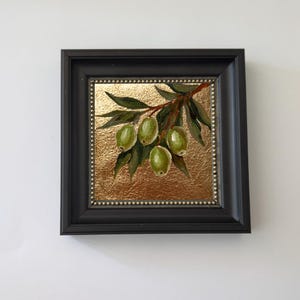 May include: A framed painting of green olives on a branch with leaves. The artwork is set against a gold background and framed in a black square frame. The painting is a close-up of the olives and leaves.