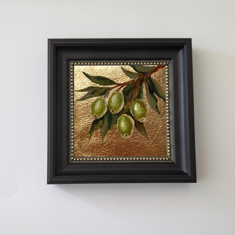 May include: A framed painting of green olives on a branch with leaves. The artwork is set against a gold background and framed in a black square frame. The painting is a close-up of the olives and leaves.