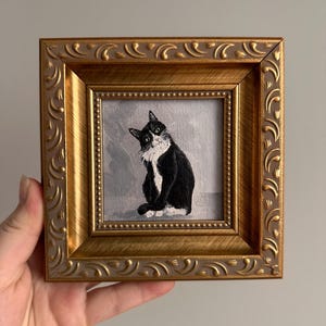 May include: A framed painting of a black and white cat with a curious expression. The cat is depicted sitting, with its head tilted. The painting is set within a gold-colored, ornate frame, adding a touch of elegance to the artwork.