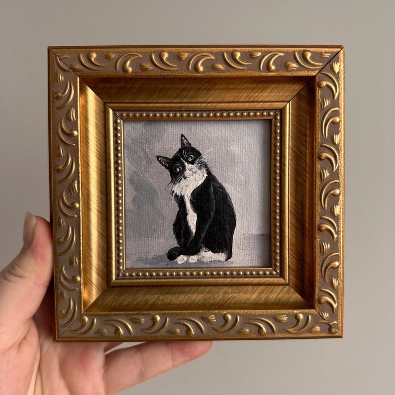 May include: A framed painting of a black and white cat with a curious expression. The cat is depicted sitting, with its head tilted. The painting is set within a gold-colored, ornate frame, adding a touch of elegance to the artwork.