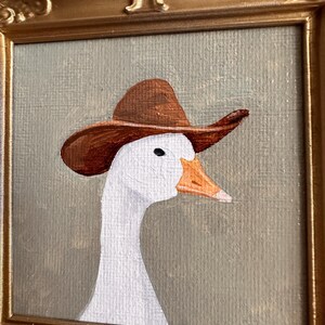 May include: A framed painting of a white goose wearing a brown cowboy hat. The goose has an orange beak and a black eye. The painting is set against a muted green background and framed in a gold-colored ornate frame.