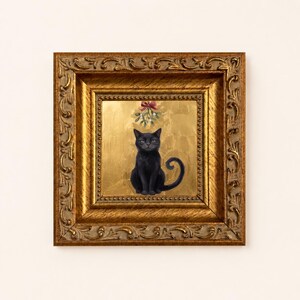 May include: A square framed painting featuring a black cat sitting centered against a textured gold leaf background. Above the cat, a sprig of mistletoe with a red bow is depicted. The artwork is encased in an ornate, antique gold-toned frame.