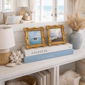May include: Two small framed paintings with gold ornate frames, depicting coastal scenes. The frames sit atop two books, one white with "COASTAL" printed on it, and one blue with "At Home". A vase with dried plants and a lamp are also visible.