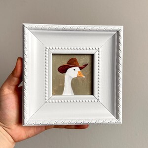 May include: A framed painting of a white goose wearing a brown cowboy hat. The artwork is set against a muted green background and framed in a white, ornate square frame. The painting is held by a hand.