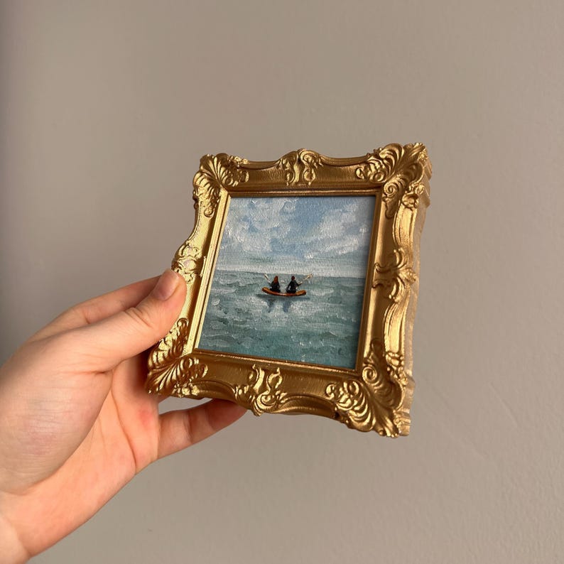 May include: A small, framed painting with a gold ornate frame. The painting depicts a seascape with a blue sky and turquoise water. Two small figures are in a boat on the water. The frame is held by a hand.