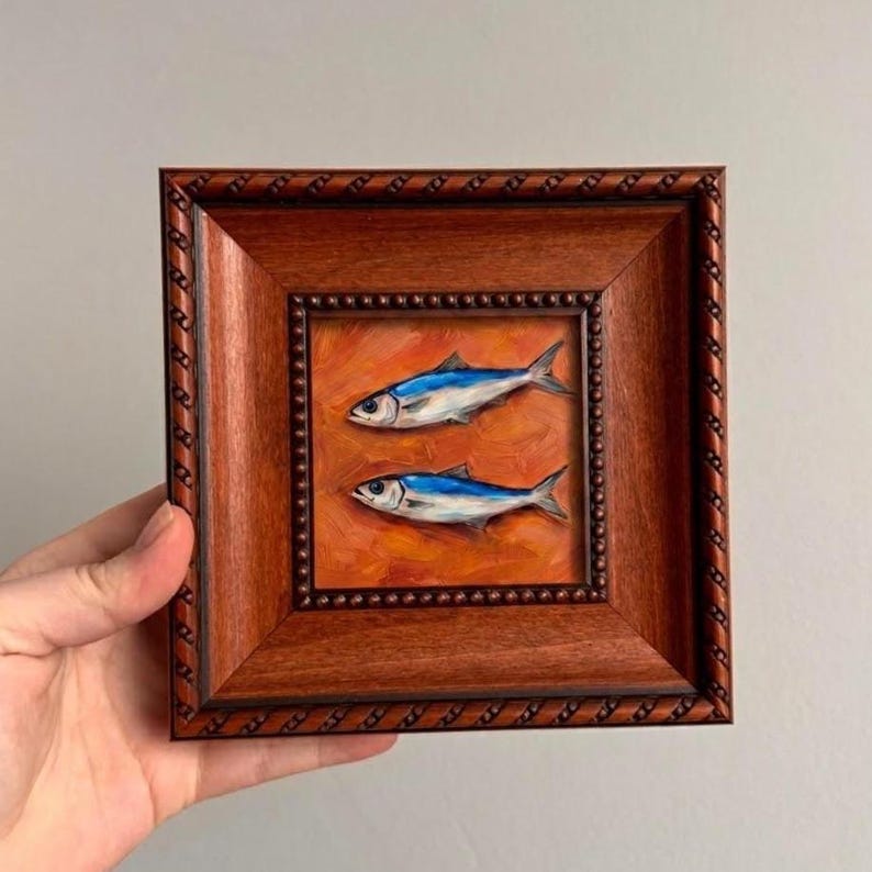 May include: A small, square, framed painting featuring two blue and silver fish on an orange background. The frame is a dark brown with a decorative rope-like border. The artwork is held in a hand.