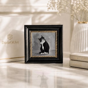 May include: Square framed artwork featuring a black and white cat. The painting is set against a gray background, framed in black with a gold border. A white box with gold lettering and a vase with flowers are in the background.