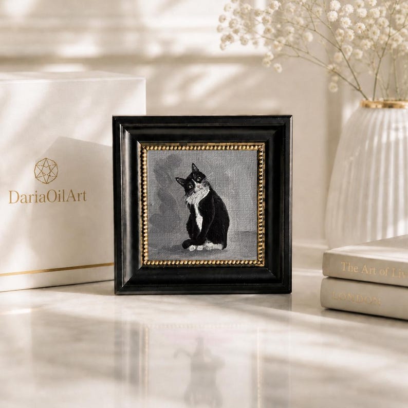 May include: Square framed artwork featuring a black and white cat. The painting is set against a gray background, framed in black with a gold border. A white box with gold lettering and a vase with flowers are in the background.