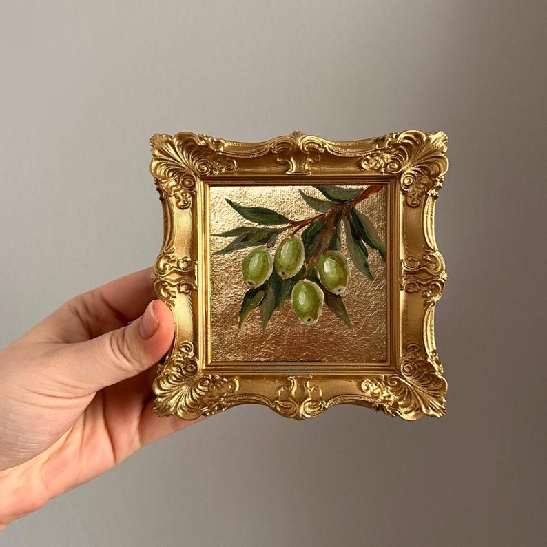 May include: A small, square, gold-colored framed painting featuring a branch of green olives and leaves. The painting has a gold background and is held by a hand.