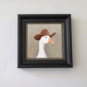 May include: Framed painting of a white goose wearing a brown cowboy hat. The artwork is set against a neutral background and framed in a black square frame with a gold beaded inner border. The goose has an orange beak.