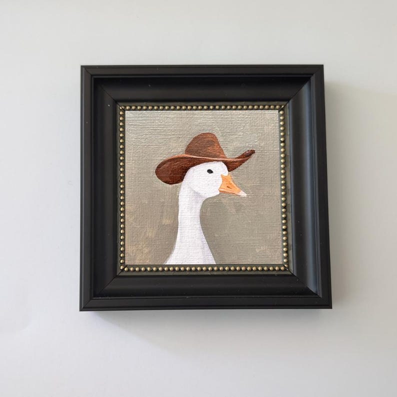May include: Framed painting of a white goose wearing a brown cowboy hat. The artwork is set against a neutral background and framed in a black square frame with a gold beaded inner border. The goose has an orange beak.