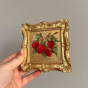 May include: A small, square painting in a gold ornate frame. The artwork depicts a cluster of vibrant red raspberries with green leaves on a branch against a gold background. The frame has a detailed, vintage design.