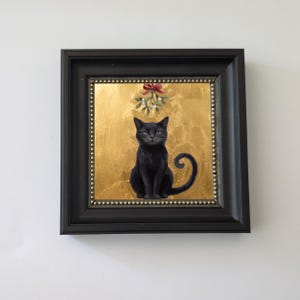 May include: A framed painting of a black cat sitting centered against a textured gold background. Above the cat hangs a sprig of mistletoe tied with a red ribbon. The artwork is enclosed in a black frame with a beaded inner border detail.