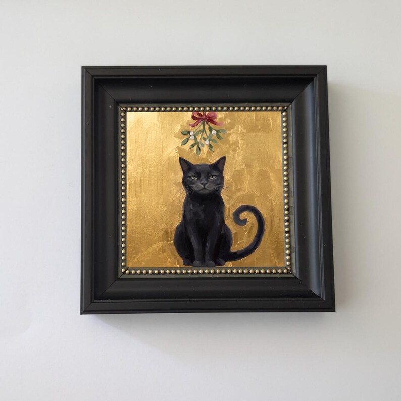 May include: A framed painting of a black cat sitting centered against a textured gold background. Above the cat hangs a sprig of mistletoe tied with a red ribbon. The artwork is enclosed in a black frame with a beaded inner border detail.