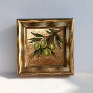 May include: A framed painting of green olives on a branch with green leaves. The artwork is set against a gold textured background and framed in a gold-toned wooden frame. The painting is a square format.