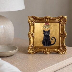 May include: A square framed artwork featuring a black cat sitting against a textured gold background. Above the cat, a sprig of mistletoe with a red bow is depicted. The frame is ornate, metallic gold, and features intricate scrollwork and carvings.