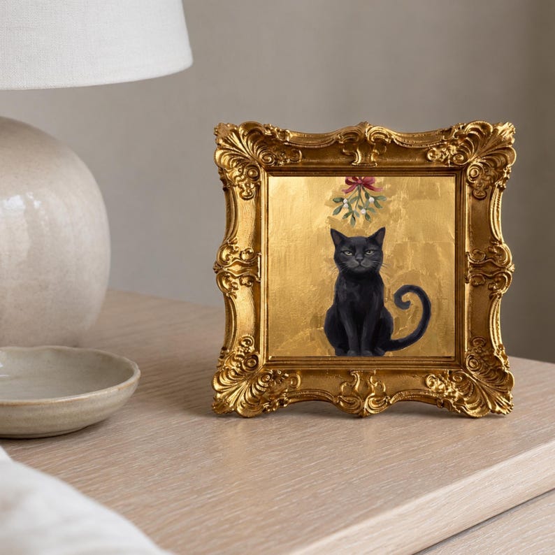 May include: A square framed artwork featuring a black cat sitting against a textured gold background. Above the cat, a sprig of mistletoe with a red bow is depicted. The frame is ornate, metallic gold, and features intricate scrollwork and carvings.
