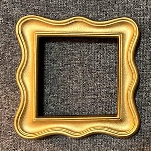 May include: A square, gold-colored picture frame with a decorative, wavy edge. The frame has a matte finish and a dark interior, creating a contrast with the bright gold exterior. The frame is empty, ready for a photo or artwork.