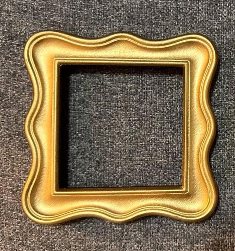 May include: A square, gold-colored picture frame with a decorative, wavy edge. The frame has a matte finish and a dark interior, creating a contrast with the bright gold exterior. The frame is empty, ready for a photo or artwork.