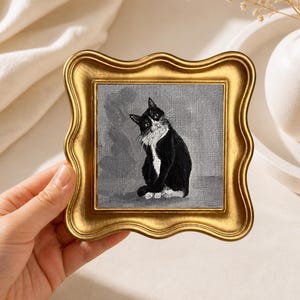 May include: A small, square painting of a black and white cat in a gold, ornate frame. The cat is sitting and looking towards the viewer. The background is a muted gray. The frame has a wavy edge.