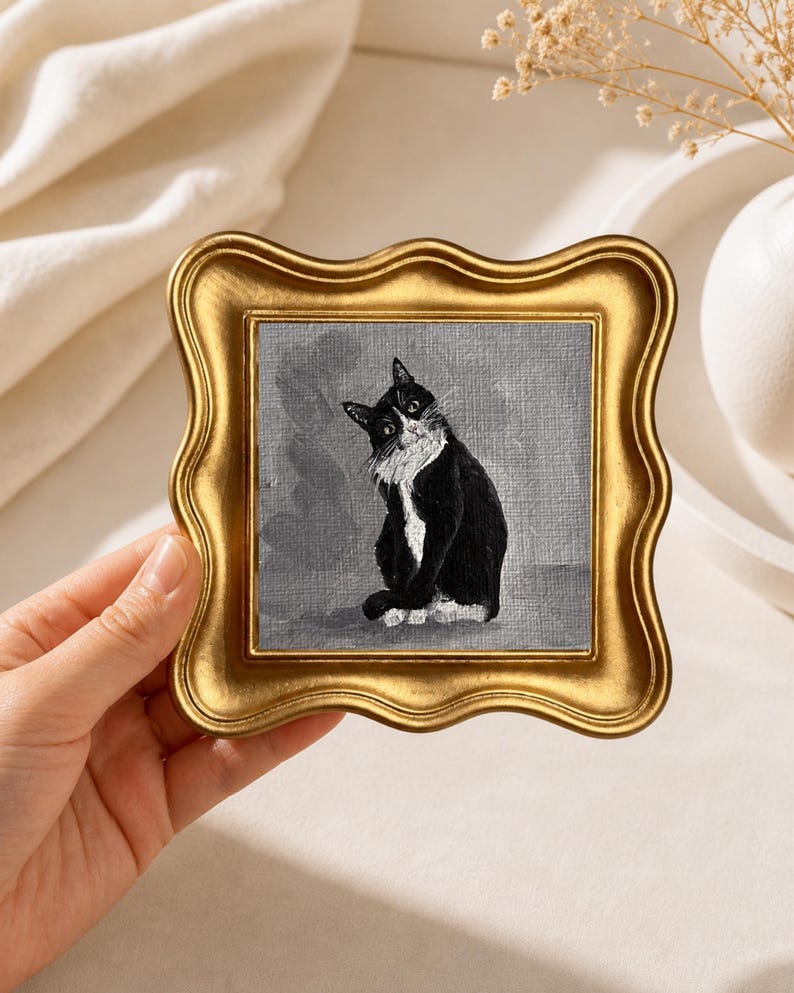 May include: A small, square painting of a black and white cat in a gold, ornate frame. The cat is sitting and looking towards the viewer. The background is a muted gray. The frame has a wavy edge.