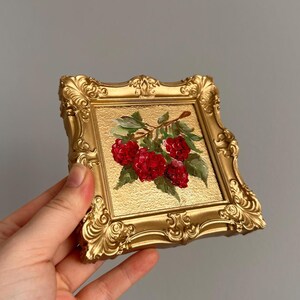 May include: A small, square, gold-colored frame with an ornate design. The framed artwork features a painting of red berries and green leaves on a gold background. The frame is held by a hand.