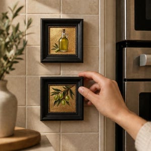 May include: Two small framed art pieces featuring olive oil and olives. The black frames have a gold textured background. One depicts a bottle of olive oil, the other a branch with green olives. The art is displayed on a tiled wall.