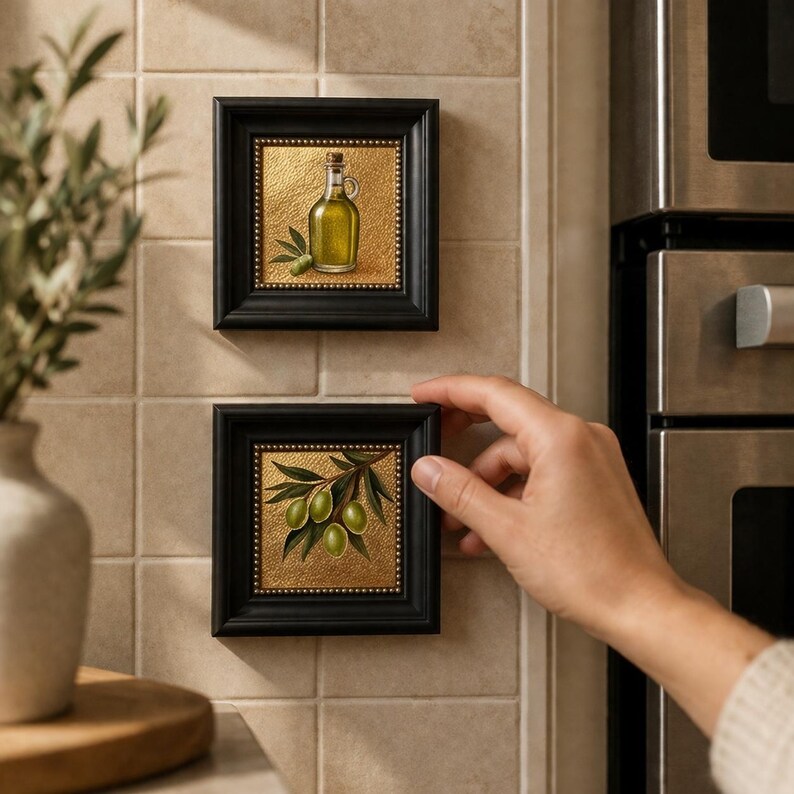 May include: Two small framed art pieces featuring olive oil and olives. The black frames have a gold textured background. One depicts a bottle of olive oil, the other a branch with green olives. The art is displayed on a tiled wall.