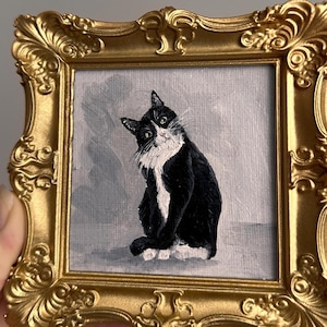 May include: A small, framed painting of a black and white cat with a gold ornate frame. The cat is depicted sitting with a curious expression against a gray background. The frame is a detailed, vintage style.