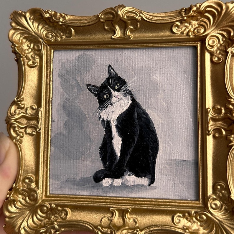 May include: A small, framed painting of a black and white cat with a gold ornate frame. The cat is depicted sitting with a curious expression against a gray background. The frame is a detailed, vintage style.
