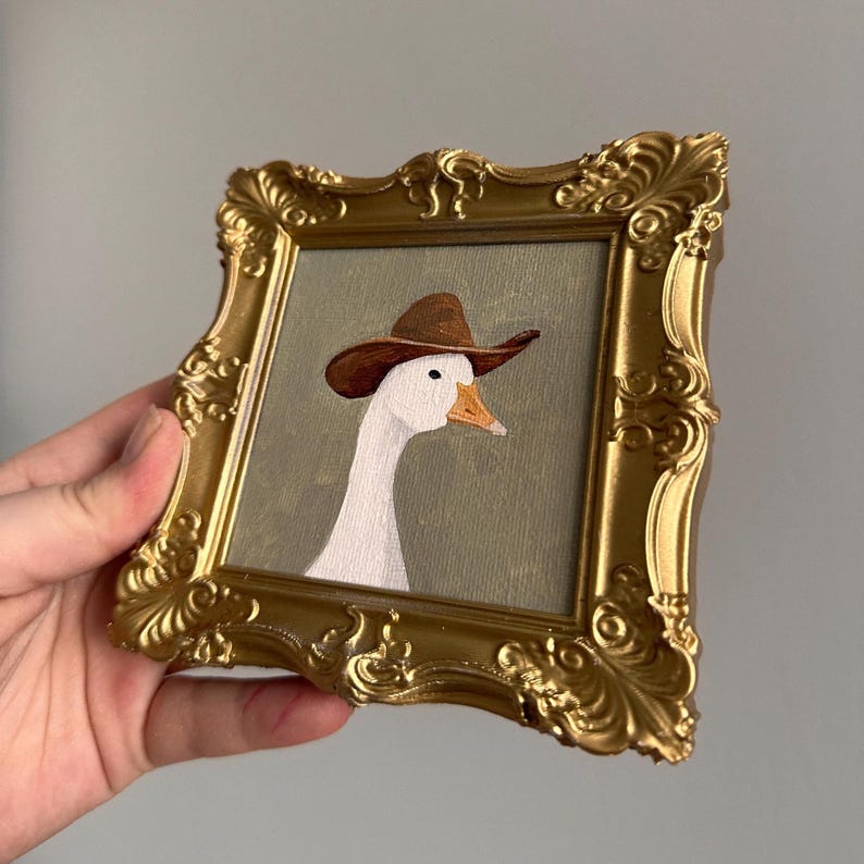 May include: A framed artwork featuring a white goose wearing a brown cowboy hat. The artwork is set against a muted green background and encased in a gold ornate frame. The image is held by a hand.