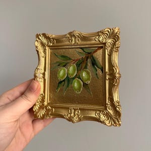 May include: A small, square painting of green olives on a branch with green leaves, set in a gold-colored ornate frame. The painting has a gold background and is held by a hand.