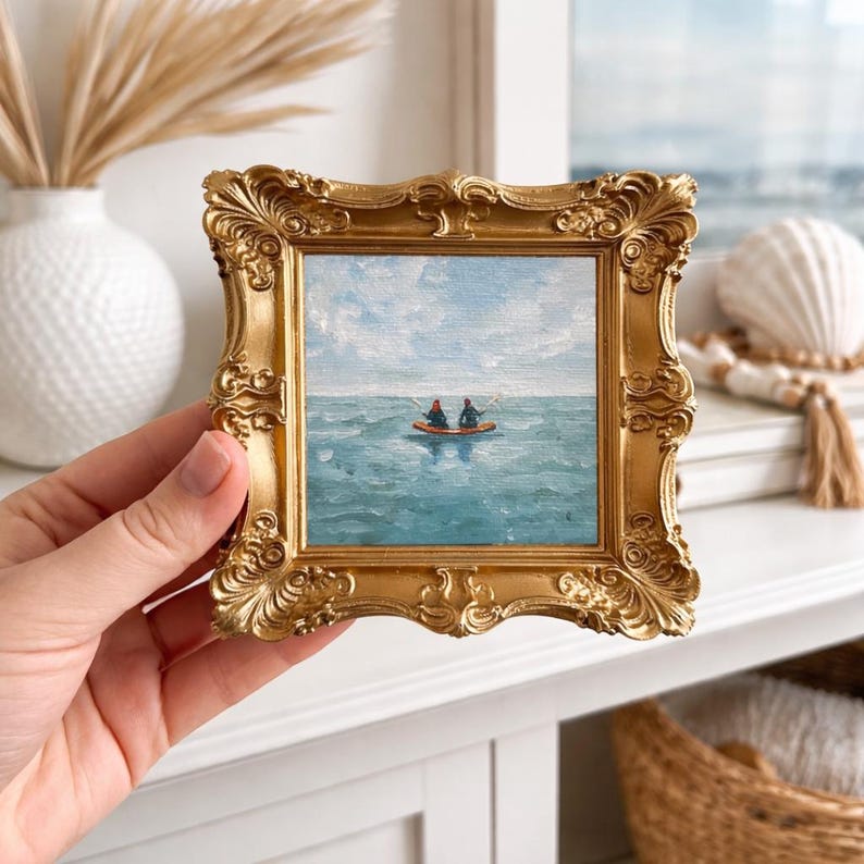 May include: A small, square painting in a gold ornate frame. The artwork depicts two figures in a boat on a blue ocean under a cloudy sky. The frame is held by a hand, with a white vase and decorative items in the background.