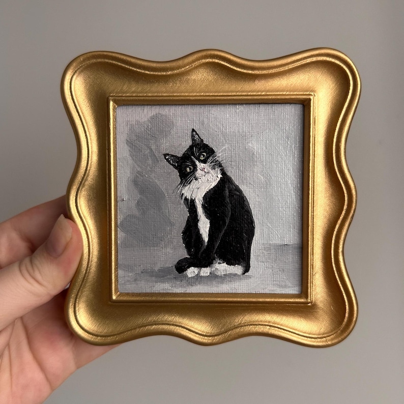 May include: A small, square painting of a black and white cat with a curious expression, set in a gold-colored, ornate frame. The cat is painted against a gray background, and the frame has a wavy, decorative edge.