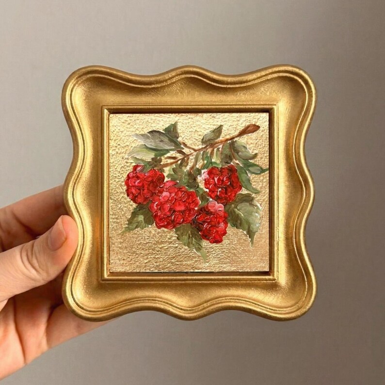 May include: A small, square, gold-framed painting featuring a cluster of vibrant red raspberries on a branch with green leaves. The background has a textured gold finish. The frame has a decorative, wavy edge.