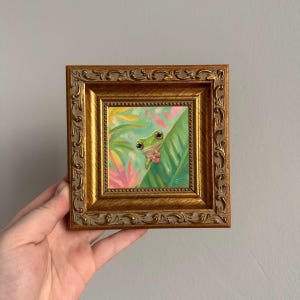 K&ouml;nnte beinhalten: Ein kleines quadratisches Gem&auml;lde eines gr&uuml;nen Frosches mit gro&szlig;en Augen, der hinter einem gro&szlig;en gr&uuml;nen Blatt hervorlugt, umgeben von rosa und gelben tropischen Blumen. Das Kunstwerk ist in einem verzierten, goldfarbenen Rahmen gefasst.