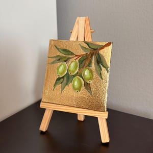 May include: A small, square painting on a gold background, depicting a branch of green olives and leaves. The artwork is displayed on a miniature wooden easel. The painting's dimensions are approximately 4 x 4 inches.