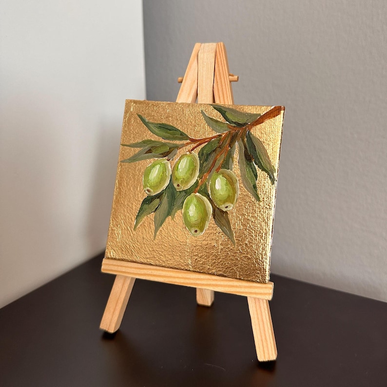 May include: A small, square painting on a gold background, depicting a branch of green olives and leaves. The artwork is displayed on a miniature wooden easel. The painting's dimensions are approximately 4 x 4 inches.