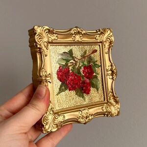 May include: A small, square, gold-colored framed painting featuring a cluster of red raspberries with green leaves on a gold textured background. The frame has ornate, detailed carvings.