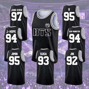 May include: A collection of black basketball jerseys featuring white trim and white lettering. The jerseys display the names and birth years of BTS members, including Jung Kook 97, J-Hope 94, Jimin 95, Suga 93, V 95, Rap Monster 94, and Jin 92.