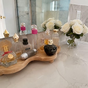 May include: A light-toned wooden tray with a curved, organic shape and small feet, holding a collection of various glass perfume bottles of different sizes and shapes, including one black bottle labeled Poison, alongside a small clear glass vase of white roses.