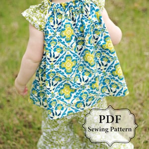 Può includere: Modello di cucito PDF per un vestito e pantaloni per bambina. Il vestito ha una stampa floreale blu e verde e i pantaloni hanno una stampa floreale verde e bianco.