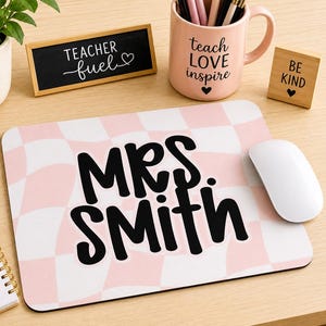 May include: A rectangular mouse pad with a light pink and white wavy checkerboard pattern featuring the black text MRS. SMITH in the center. The mouse pad is positioned on a desk surface next to a white computer mouse, a pink mug, and a notebook.