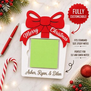 May include: A white wooden note holder shaped like a gift with a large red bow at the top. The bow features the text Merry Christmas. The center holds a bright green square sticky note. The bottom is personalized with the names Asher, Ryan, & Dilan.