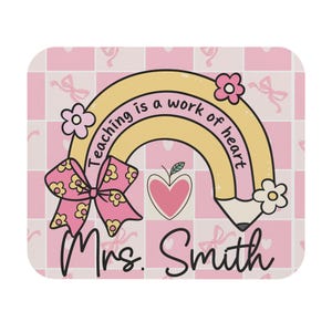 Può includere: Un tappetino per mouse rettangolare con sfondo a scacchi rosa chiaro e bianco con piccoli fiocchi rosa. Il design include una matita gialla a forma di arcobaleno con il testo Teaching is a work of heart, un cuore rosa e Mrs. Smith.