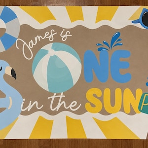 May include: A rectangular poster featuring a yellow and white sunburst background with a brown center. It displays the text "James is ONE in the SUN" and "POOL" on a sign. Graphics include a blue flamingo float, a beach ball, a life ring, and sunglasses.