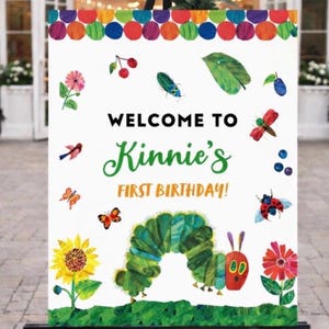 May include: A white sign with colorful polka dots, flowers, and insects, reads "WELCOME TO Kinnie's FIRST BIRTHDAY!". A large green caterpillar is featured at the bottom. The sign is displayed on a black easel.