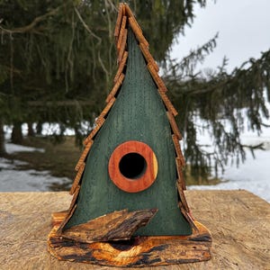 Könnte beinhalten: Handgefertigtes Vogelhaus aus Holz mit grünem Korpus und einem Dach aus Naturholz. Das Vogelhaus hat ein rundes Eingangsloch mit Holzverkleidung und einen rustikalen Holzsockel. Das Design erinnert an eine kleine Hütte.