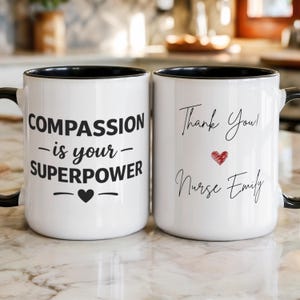 Pode incluir: Duas canecas de cerâmica brancas com bordas e alças pretas. Uma caneca diz "COMPASSION - is your - SUPERPOWER" com um coração. A outra caneca diz "Thank You! Nurse Emily" com um coração vermelho.